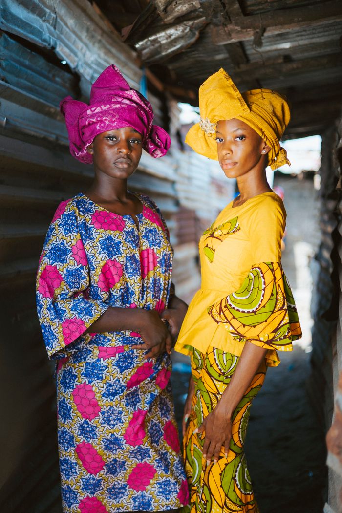 Two women in vibrant traditional attire pose in an alley in Monrovia, showcasing cultural fashion.